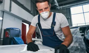 Mechanik w masce i kombinezonie szlifuje maskę samochodu.