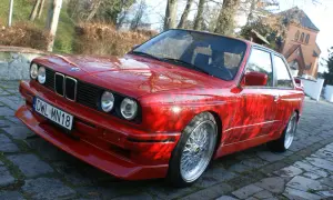 Czerwone BMW E30 z dokładanym spoilerem IS, na tle drzew i budynku.