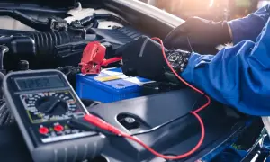 Mechanik w rękawiczkach podłącza prostownik do akumulatora samochodowego, obok leży multimetr.