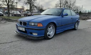 Niebieskie BMW E36 coupe z czarnym spoilerem na przednim zderzaku, zaparkowane na zewnątrz.