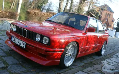 Czerwone BMW E30 z dokładanym spoilerem IS, na tle drzew i budynku.