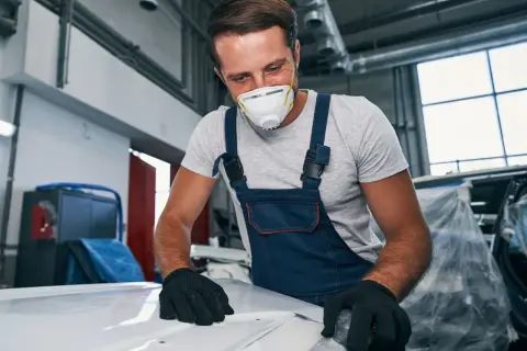 Mechanik w masce i kombinezonie szlifuje maskę samochodu.