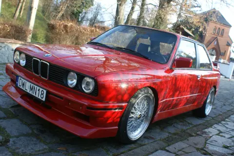 Czerwone BMW E30 z dokładanym spoilerem IS, na tle drzew i budynku.
