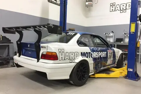 BMW E36 coupe z dużym spojlerem na podnośniku w warsztacie.