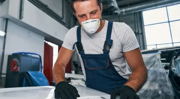 Mechanik w masce i kombinezonie szlifuje maskę samochodu.