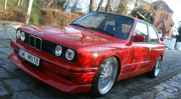Czerwone BMW E30 z dokładanym spoilerem IS, na tle drzew i budynku.