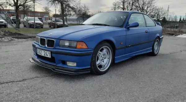 Niebieskie BMW E36 coupe z czarnym spoilerem na przednim zderzaku, zaparkowane na zewnątrz.