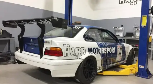 BMW E36 coupe z dużym spojlerem na podnośniku w warsztacie.
