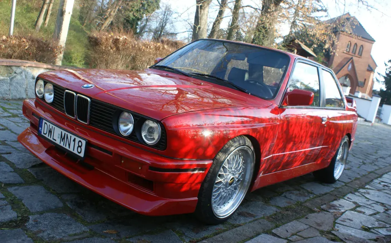 Czerwone BMW E30 z dokładanym spoilerem IS, na tle drzew i budynku.