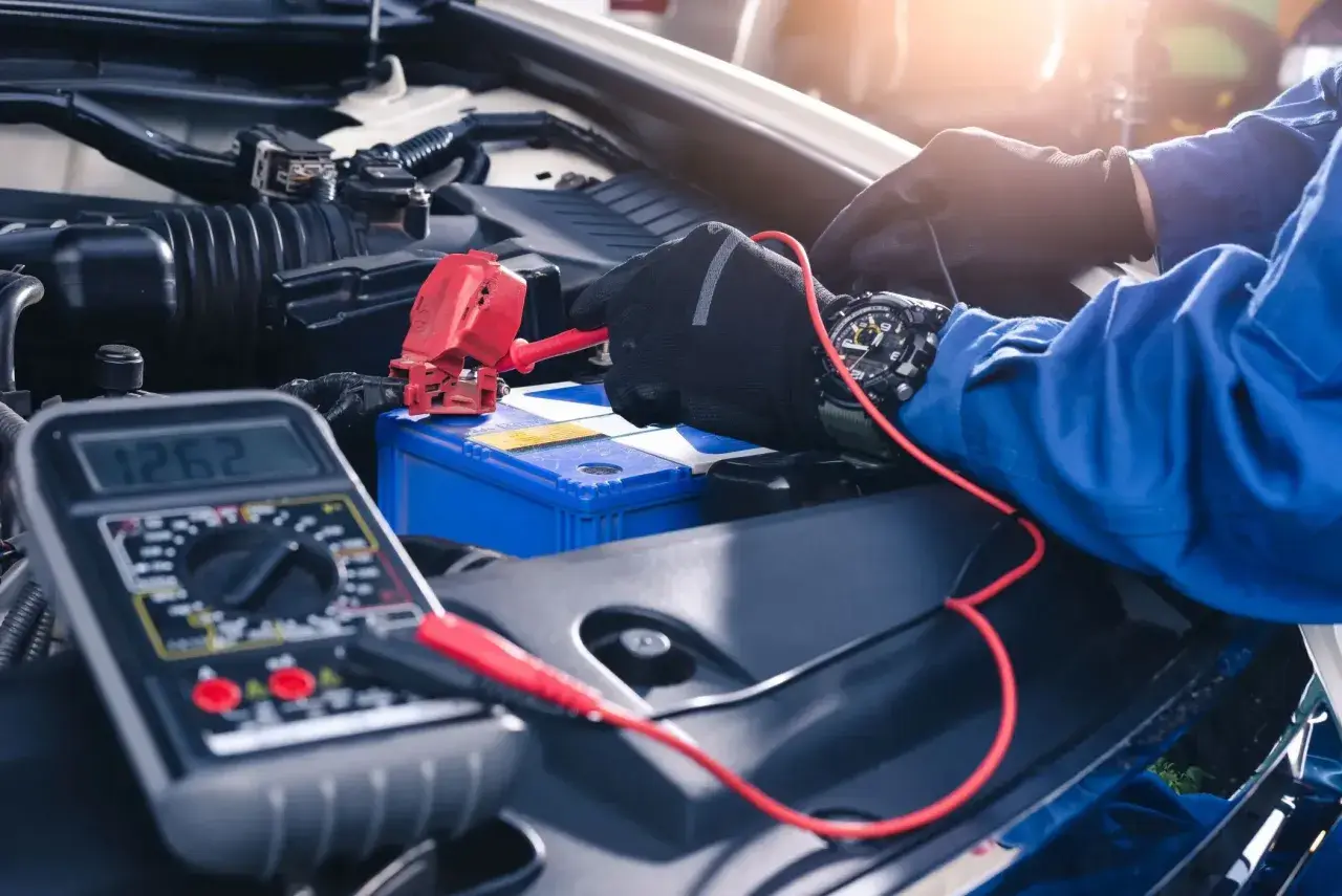Mechanik w rękawiczkach podłącza prostownik do akumulatora samochodowego, obok leży multimetr.
