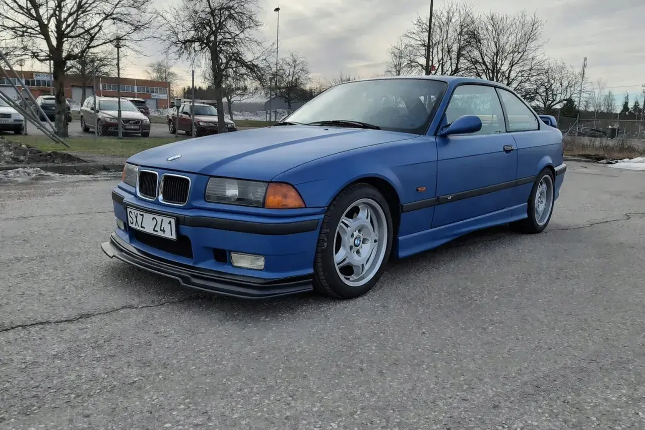 Niebieskie BMW E36 coupe z czarnym spoilerem na przednim zderzaku, zaparkowane na zewnątrz.