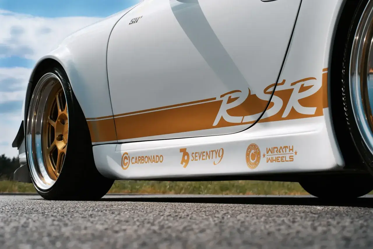 Biały samochód z pomarańczowymi naklejkami "RSR", "CARBONADO", "SEVENTY9" i "WRATH WHEELS".