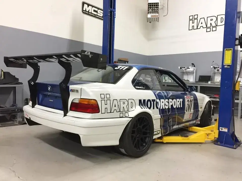 BMW E36 coupe z dużym spojlerem na podnośniku w warsztacie.