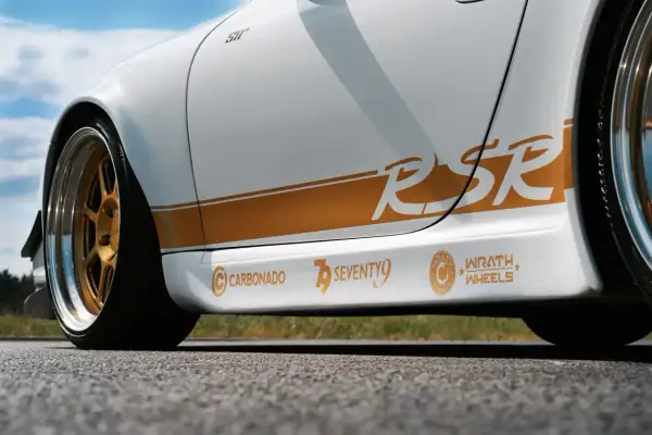 Biały samochód z pomarańczowymi naklejkami "RSR", "CARBONADO", "SEVENTY9" i "WRATH WHEELS".