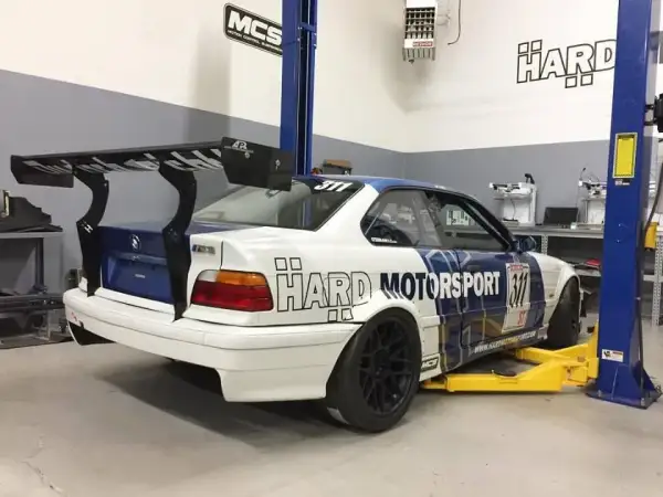 BMW E36 coupe z dużym spojlerem na podnośniku w warsztacie.